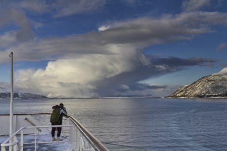 Eine Person fotografiert von einem Schiff aus die Natur aus. Man sieht das Meer, ein weißes Wolkengebilde und vereinzelt schneebedeckte Berge.