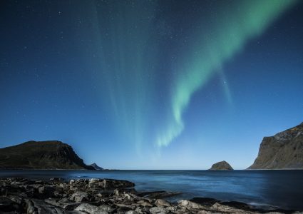 Ein Blick auf die grünen Nordlichter am blauen Sternenhimmel. Man sieht einen setinigen Boden der ins Meer führt und im Hintergrund sind einzele Berge zu sehen.